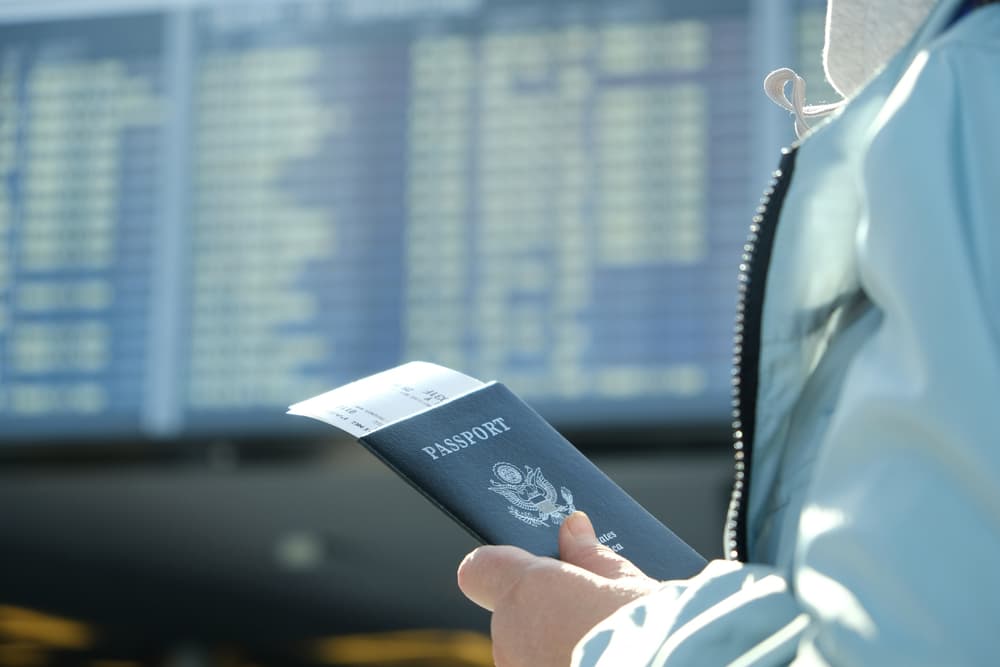 Hand holding passport in front of board with flight schedules.