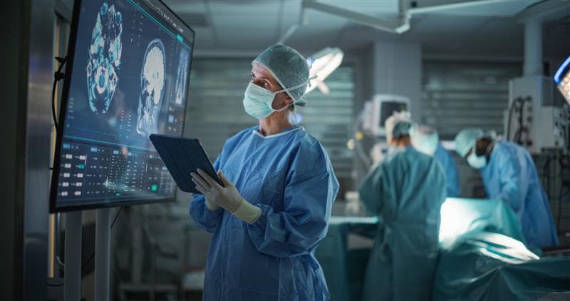 Surgeon Reviews Critical Brain Scans Displayed on a Monitor as Well as Health Data on a Tablet Computer. Surgeon Reviews Critical Brain Scans Displayed on a Monitor as Well as Health Data on a Tablet Computer.
