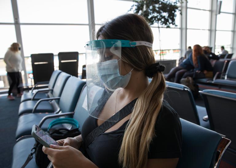 Young woman in airport terminal wearing PPE and social distancing regardless of vaccination status.